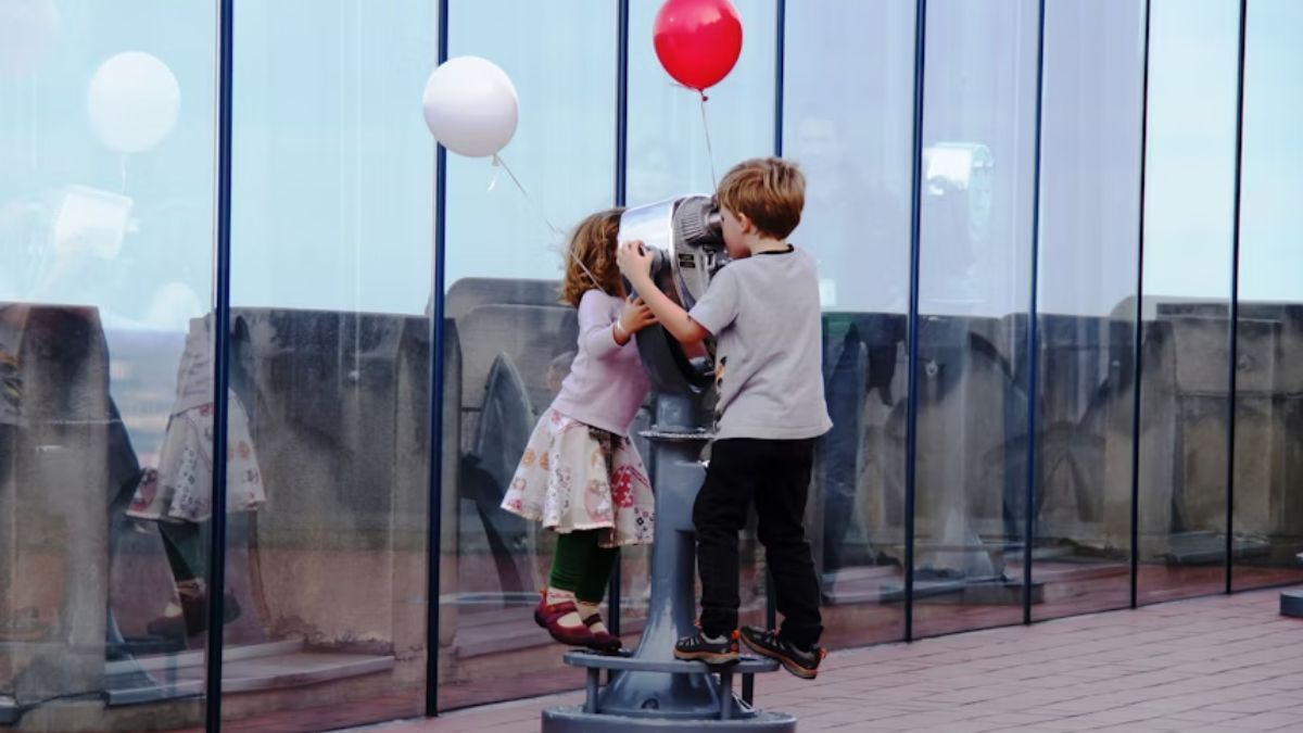 A boy and a girl playing near a parking meter beside a steel-and-glass building in the soft afternoon light, with the street in gentle focus.