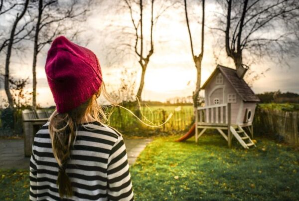 A girl wearing a red beanie walks through a sunlit meadow toward a house framed by the glowing sun.