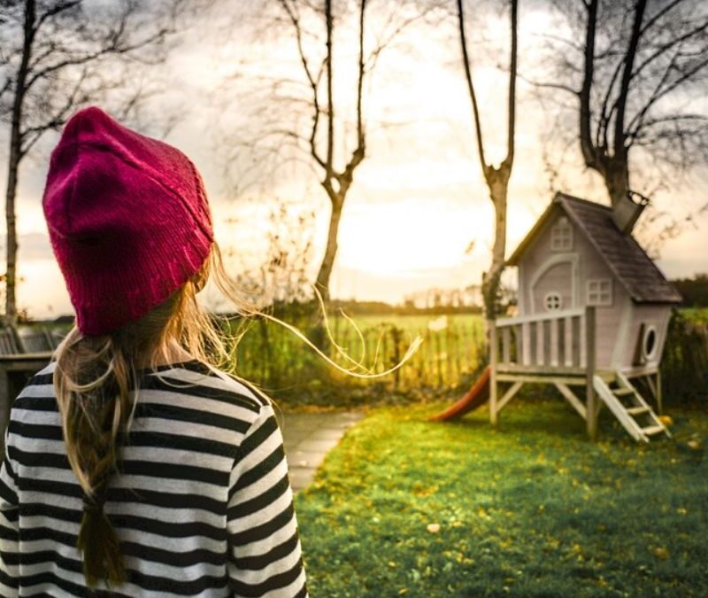 A girl wearing a red beanie walks through a sunlit meadow toward a house framed by the glowing sun.