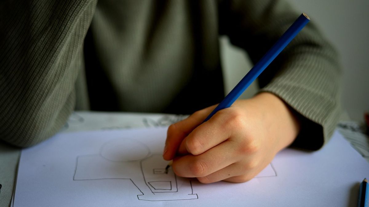 Only the hands of a young boy are visible, holding a pen and drawing in a sketchbook for an assessment.