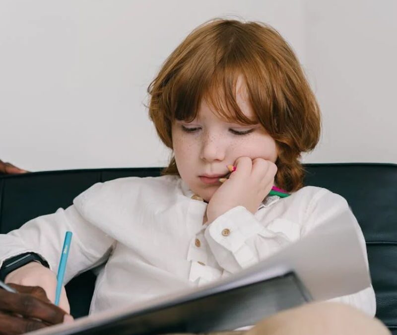 A young boy holding a notebook while sitting on a couch, guided by a therapist.
