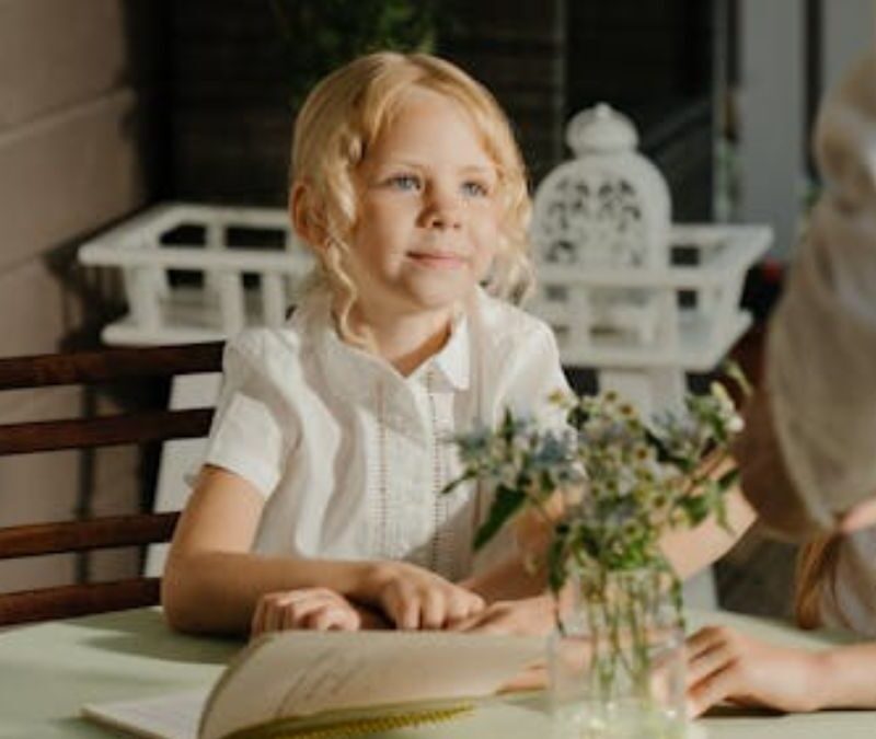 An opened book with a blonde-haired child looking expectant in a sunlit room.
