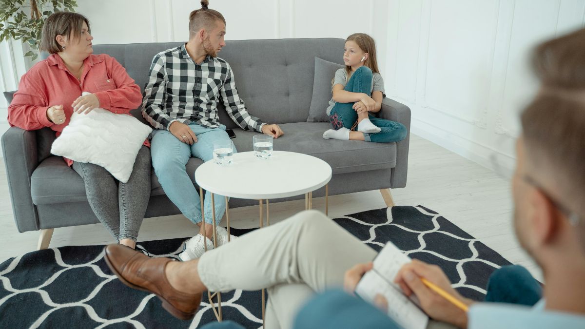 Parents sit in a therapy room with their child while the therapist observes with legs crossed.