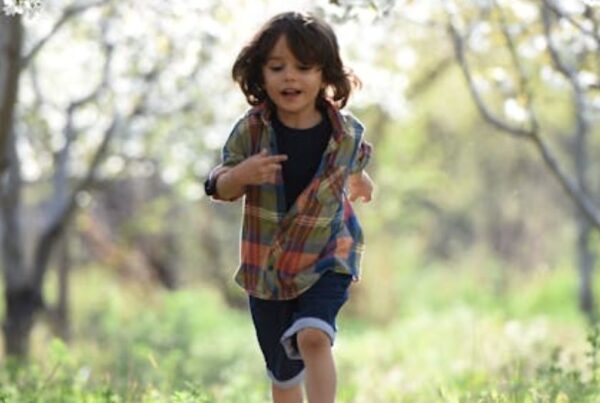 A boy running out of a sunlit forest into a meadow, wearing a plain shirt and looking happy and energetic.