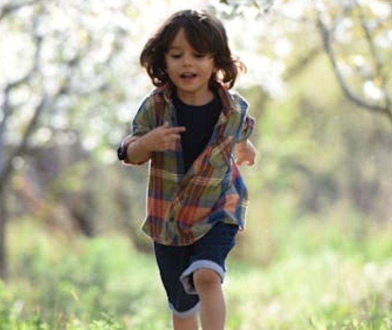 A boy running out of a sunlit forest into a meadow, wearing a plain shirt and looking happy and energetic.