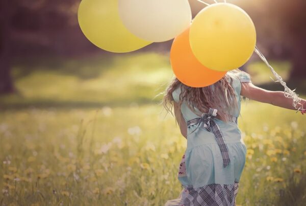 A young girl runs across a meadow holding balloons, her back to the camera, illustrating freedom and play in daily life for a child with autism.