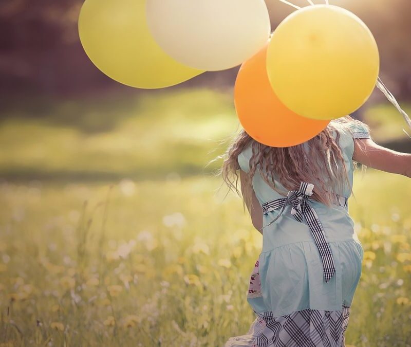 A young girl runs across a meadow holding balloons, her back to the camera, illustrating freedom and play in daily life for a child with autism.