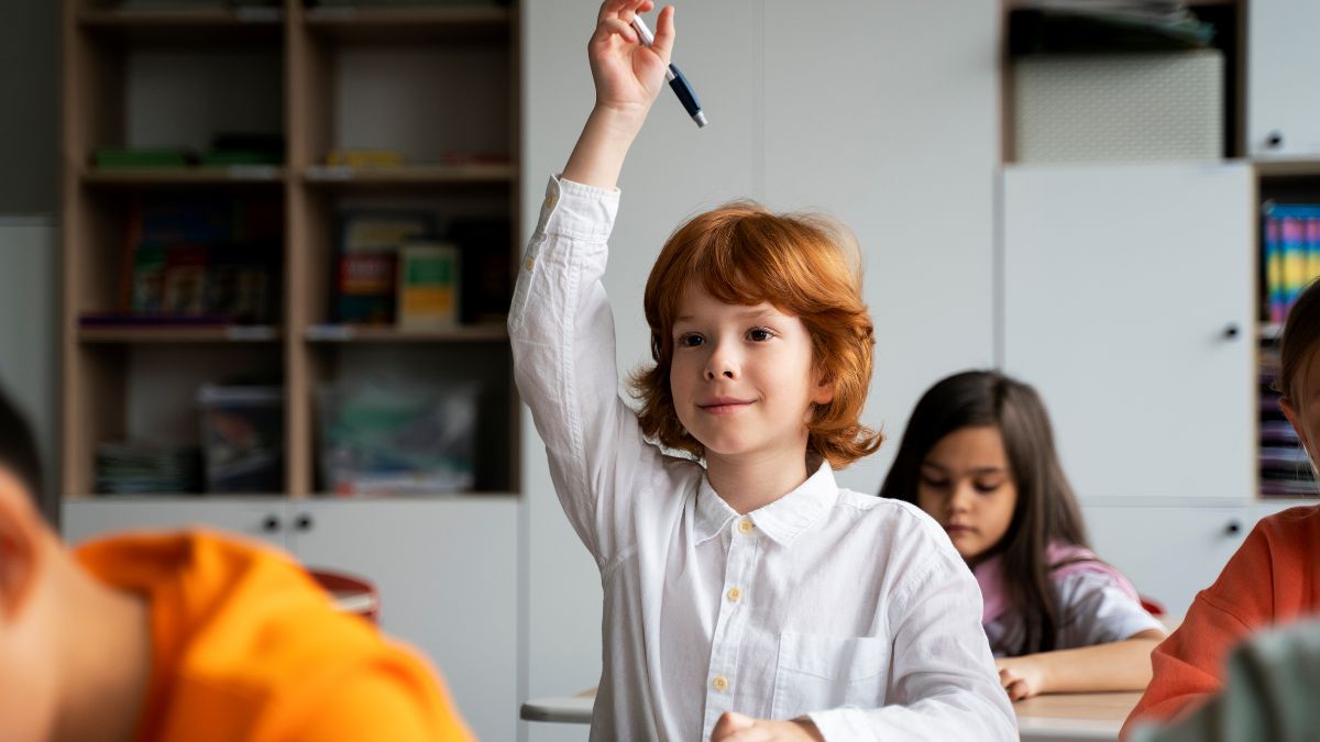 A red-haired boy in a white shirt raises his hand confidently in class, ready to recite his answer.