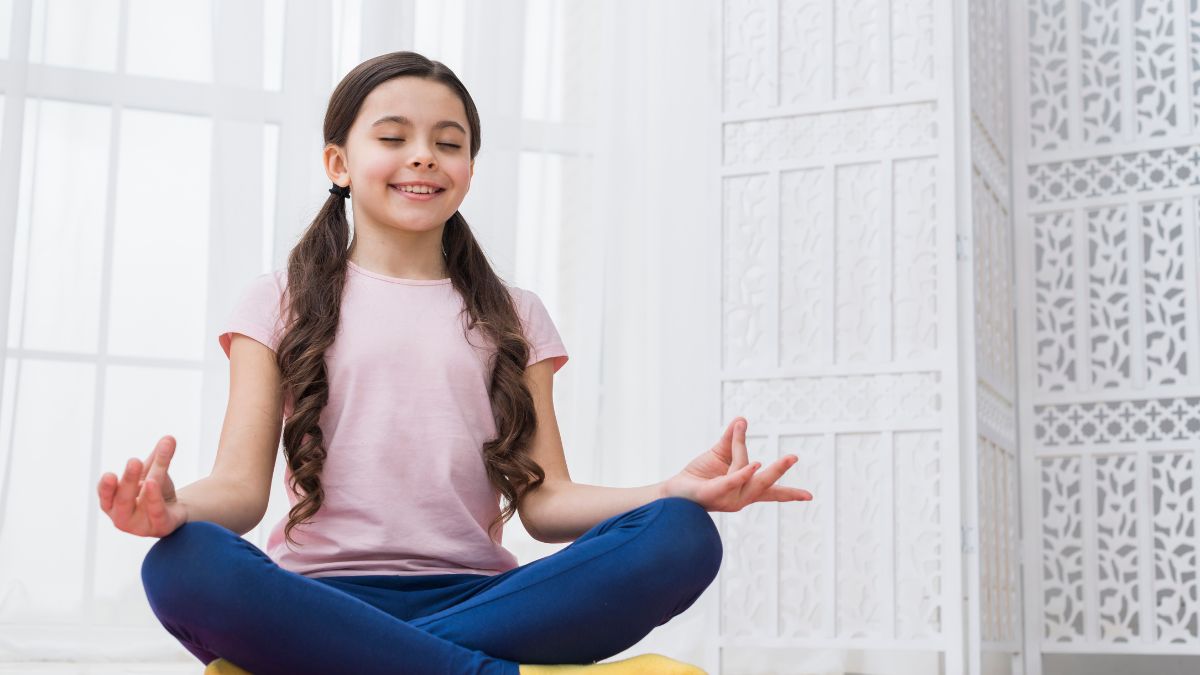 A young girl with autism in a pink blouse sits cross-legged in a brightly lit white room, meditating to cope with overstimulation.