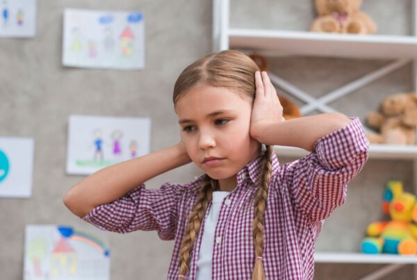 A young girl with autism wearing a pink blouse covers her ears in an empty schoolroom, illustrating overstimulation.