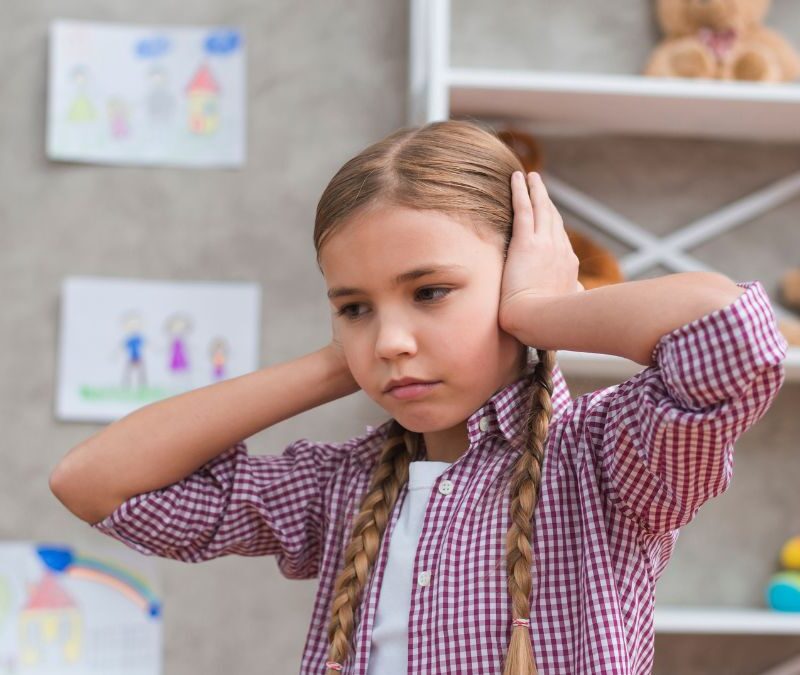 A young girl with autism wearing a pink blouse covers her ears in an empty schoolroom, illustrating overstimulation.