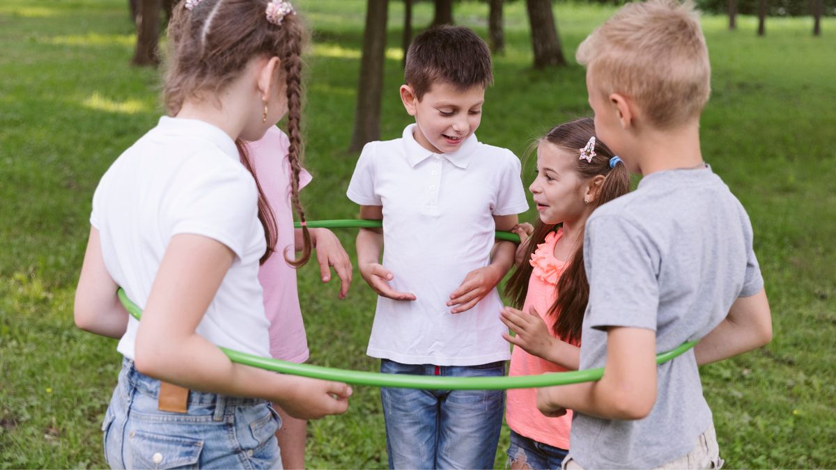 A group of children plays outdoors, gathering together inside a large hula hoop on a grassy lawn, participating in a fun, cooperative activity during a behavioral therapy session.