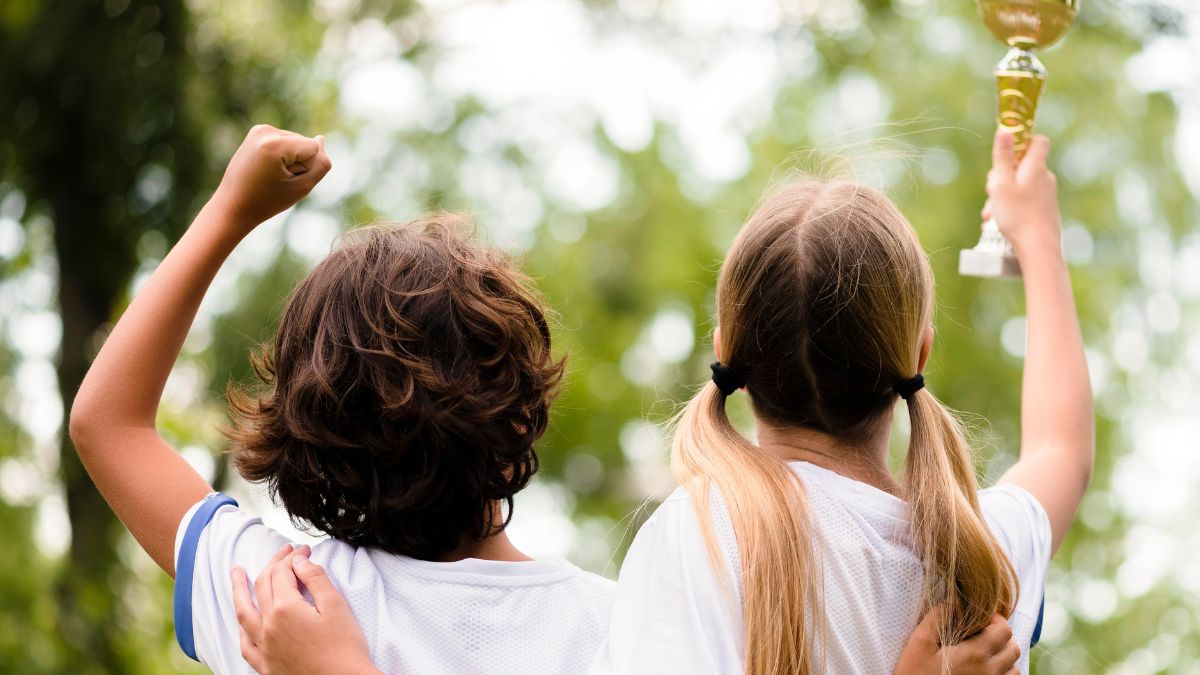A pair of children wearing soccer shirts stand with their backs to the camera, raising their hands triumphantly after winning an outdoor game in a field during an ABA therapy session.