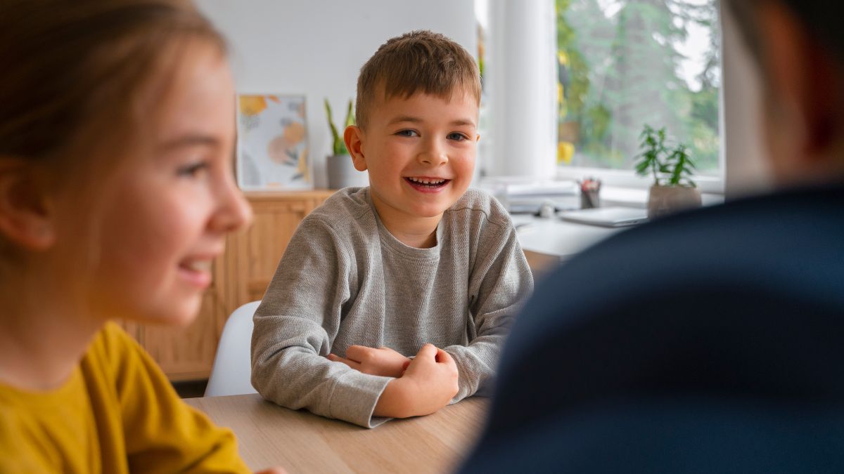 A group of children exchange friendly greetings in a brightly lit classroom, with an open window showing trees and foliage outside.