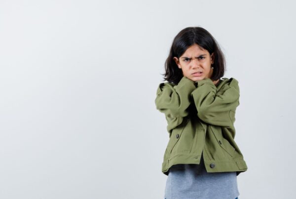 A young girl in a green shirt stands against a gray background with a frowning expression, appearing upset.