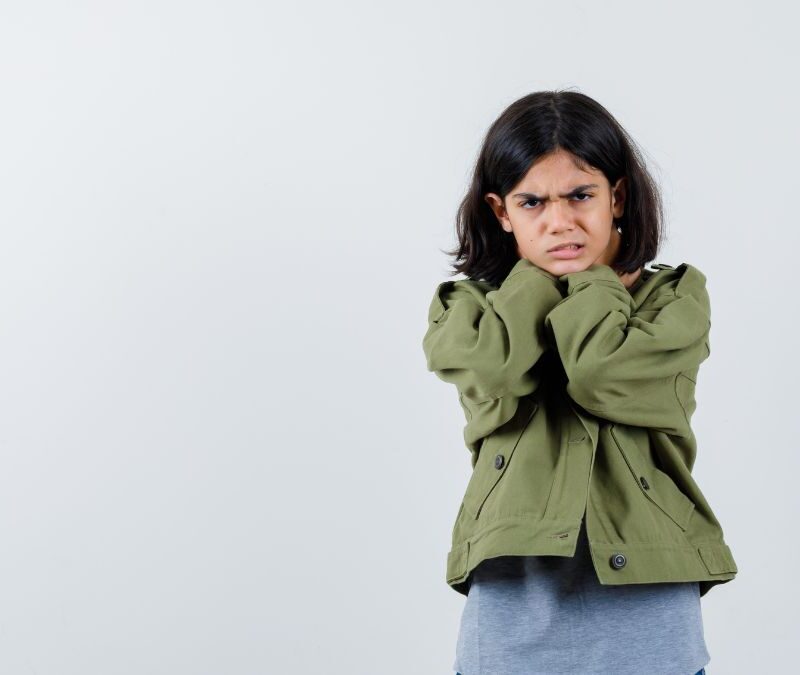 A young girl in a green shirt stands against a gray background with a frowning expression, appearing upset.