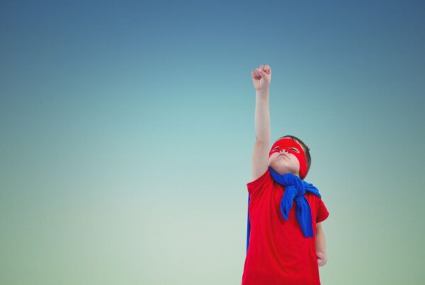 A young boy wearing a superhero costume poses against a dark blue background, stretching his arm with a folded fist skyward as if preparing to fly during an ABA therapy-themed activity.