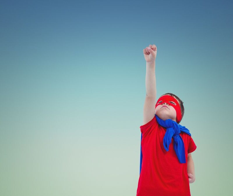 A young boy wearing a superhero costume poses against a dark blue background, stretching his arm with a folded fist skyward as if preparing to fly during an ABA therapy-themed activity.