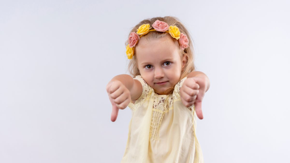 A young girl wearing a flower headband looks at the camera while signing a thumbs-down during functional communication training.